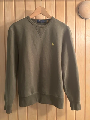 Sweatshirt från Polo Ralph Lauren - Olivgrön sweatshirt från Polo Ralph Lauren i superfint skick med klassisk rund hals och gul broderad logga på bröstet. Tröjan har ribbade muddar vid ärmslut och nederkant samt långärmad passform.