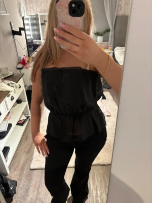 Svart bandeau tubtopp från Bikbok - Säljer en svart bandeau tubtopp från Bikbok i storlek L. Toppen har en glansig satinliknande yta med lätt struktur, är axelbandslös och har en snygg volang nedtill. Perfekt för dig som gillar stilrena och trendiga plagg med lite extra edge.