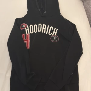 Svart hoodie från Hoodrich med patchar - Svart hoodie från Hoodrich i storlek L med vita och röda broderade patchar och tryck på bröstet. Tröjan har huva med snörning och ribbade muddar. Snygga detaljer som Hoodrich-märke på ärmen och stor broderad siffra 4. Perfekt för streetwear-stilen.