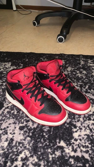 Nike Air Jordan 1 Mid röd/svart 39 - Säljer ett par Nike Air Jordan 1 Mid i klassisk röd och svart färg med vita sulor. Skorna har svart swoosh, snörning och perforerad tå. Tillverkade i skinn och textil, ikonisk Jordan-logga på plösen. Perfekt för dig som vill sticka ut med en tidlös sneaker, små skador som knappt syns o liten vit fläck bakom ena skon.