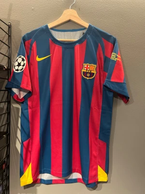 Barcelona Ronaldinho 10 fotbollströja - Säljer en officiell FC Barcelona fotbollströja från Nike med Ronaldinho och nummer 10 på ryggen. Tröjan har breda blå och röda ränder, gult Nike-märke och klubbemblem på bröstet. Champions League-märke på ärmen och detaljer i gult. Tillverkad i lätt polyester. Helt ny tröja en replika med tagg.