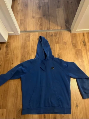 Blå hoodie från Under Armour - Säljer en blå hoodie från Under Armour i storlek L. Tröjan har huva med snörning, en stor magficka och långärmade raglanärmar. Materialet är mjukt och känns fleeceaktigt på insidan, perfekt för chill eller träning. UA-logga på bröstet för extra stil.Passsar med fördel någon i storlek medium tröjan är i bra bruksskick.