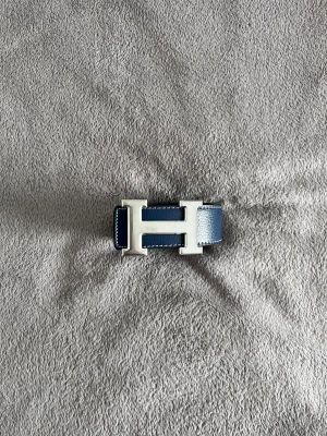 Hermès bälte  - Säljer ett stilrent Hermès bälte med den ikoniska H-spännet i silver. Ena sidan är marinblå och andra sidan är svart, båda i grymt snyggt skinn med vit kontrastsöm. Perfekt för dig som vill ha en lyxig accessoar med clean look och möjlighet att variera färg.