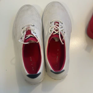 Vita sneakers från Tommy Hilfiger - Snygga vita sneakers från Tommy Hilfiger med klassisk design. Skorna har vita snören, röd insida och detaljer i marinblått samt logga på hälen. Tillverkade i textil med platt sula och rund tå. Perfekta för en clean och sportig stil.