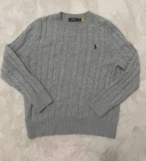 Ralph Lauren tröja - Superfin och klassisk kabelstickad tröja i ljusgrått. Mjuk, varm och sitter snyggt. Perfekt för både vardag och lite finare tillfällen.  ✔ Äkta Polo Ralph Lauren ✔ Ikoniska ryttaremblemet ✔ Bra skick – inga fläckar eller hål  Storlek: S Passform: Normal Färg: Ljusgrå