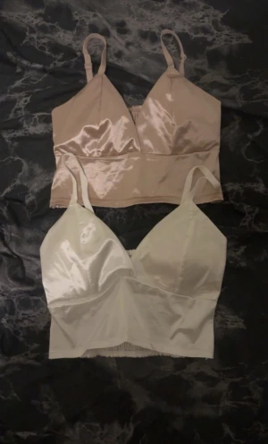 Champagne och vit satin linnetopp - Två stilrena linnetoppar i satin, en i champagne och en i vit. Båda har justerbara axelband och en croppad passform. Framsidan är slät och glansig medan baksidan har ribbad, smockad struktur för extra komfort och snygg look. Perfekt för att lyfta din outfit.