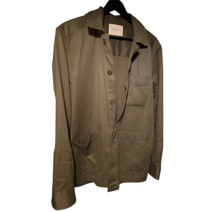 Khaki overshirt från These Glory Days - Snygg beige overshirt från These Glory Days med klassisk krage, tre framfickor och knäppning med knappar. Jackan är tillverkad i mjuk bomull och har en clean, minimalistisk look som passar perfekt till jeans eller chinos. Har en fläck som antagligen tas bort i tvätten.