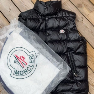 Moncler Väst - Snygg svart dunväst från Moncler med glansig finish och klassisk logga på bröstet. Västen har hög krage, dragkedja i sidan och tryckknappar framtill. Perfekt för lager-på-lager och ger en clean, sportig look. Size 3=L men sitter bra på M med beror lite på längd och vikt(passar mig som har M i vanliga fall) Dustbag och tags följer med även kvitto