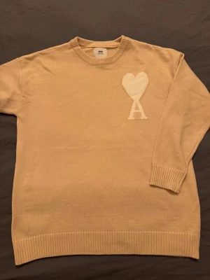 Beige stickad tröja från Ami - Säljer en beige stickad tröja från Ami Alexandre Mattiussi med ett vitt hjärta och bokstaven A på bröstet. Tröjan har rund hals, ribbade muddar och lång ärm. Perfekt för dig som gillar stilrena detaljer och mjuka material.