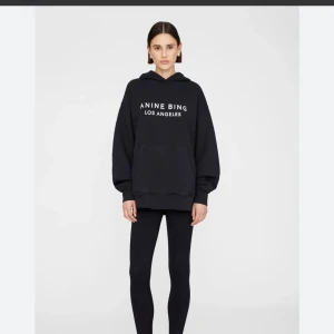 Svart hoodie från Anine Bing - Svart oversized hoodie från Anine Bing med vit text 'ANINE BING LOS ANGELES' på bröstet. Tröjan har huva, stor magficka och långa ärmar med ribbade muddar. Perfekt för en chill och cool look.