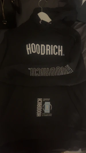 Svart hoodie från Hoodrich med tryck - Svart hoodie från Hoodrich i storlek M med stor vit logotyp på bröstet och detaljerat tryck på ryggen. Tröjan har huva med snörning och coola grafiska prints i vitt och ljusblått. Perfekt för dig som gillar streetwear och vill sticka ut.