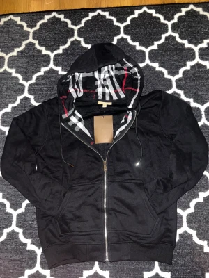 Svart hoodie från Burberry med dragkedja - Svart hoodie från Burberry med klassiskt rutigt foder i huvan i svart, vitt och rött. Tröjan har broderad Burberry-logga på bröstet, snörning i huvan och praktiska fickor framtill. Materialet är mjuk bomull och passformen är normal.