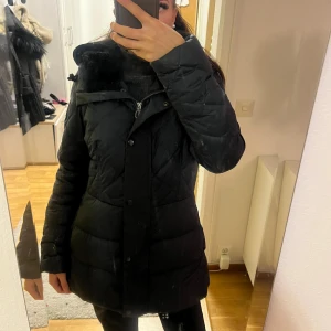 Svart dunjacka från G-Star RAW - Svart dunjacka från G-Star RAW med quiltad design, huva och mysig fuskpälsdetalj vid kragen. Jackan har dragkedja och knappar framtill, samt en figurnära passform som ger en snygg siluett. Perfekt för kalla dagar.