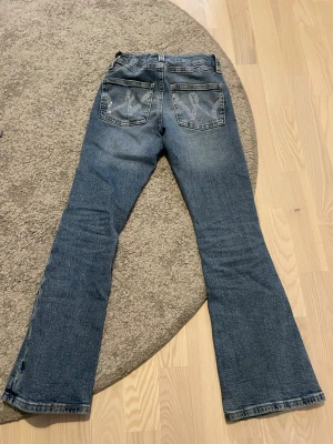 Blå bootcut jeans med strassdetaljer - Snygga blå jeans med bootcut passform och coola strassdetaljer på bakfickorna. Jeansen har klassisk femficksdesign, knapp och dragkedja framtill samt lätt slitning för en avslappnad vibe. Perfekta för dig som vill sticka ut lite extra.