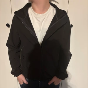 Svart vindjacka från Colmar - Svart vindjacka från Colmar Originals med huva och dragkedja framtill. Jackan har en stilren look, Colmar-logga på ärmen och praktiska fickor med dragkedja. Perfekt för dig som gillar sportig och clean stil. Grisch, Laidback! 