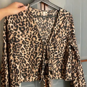 Leopardmönstrad omlottblus från NELLY - Säljer en croppad omlottblus från NELLY med leopardmönster i beige och svart. Blusen har långa, vida ärmar och knyt framtill som ger en snygg v-ringad look. Perfekt för dig som gillar att sticka ut med djurmönster och coola detaljer.