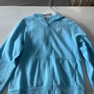 Ljusblå Nike hoodie med dragkedja - Nike hoodie i ljusblått med broderad vit logga på bröstet. Tröjan har huva, hel dragkedja framtill och två stora fickor. Mjuk insida och klassisk streetstil – perfekt för chill dagar eller träning.