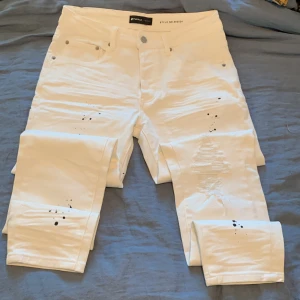 Vita jeans från Purple Brand med detaljer - Säljer ett par vita jeans från Purple Brand, modell 002024. Byxorna har svart färgstänk, slitningar på framsidan och klassisk femficksdesign. De är raka i modellen och har en snygg, modern look med coola detaljer. Perfekta för dig som vill sticka ut.
