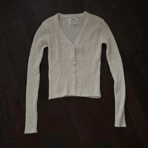 Beige ribbad kofta från Zara - Säljer en beige ribbad kofta från Zara med v-ringning och tre knappar framtill. Koftan har långärmade smala ärmar, är tajt och är tillverkad i ett mjukt stickat material. Perfekt att slänga över en topp för en chill look. Tyvärr har jag klippt av tvättlappen men den tvättas i 30😊