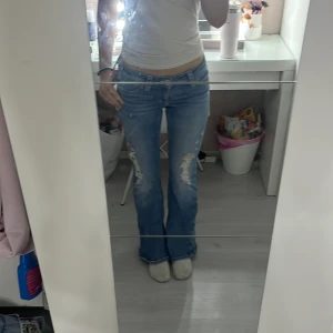 Blå bootcut jeans från Gina Tricot - Snygga blå low waist jeans från Gina Tricot i storlek S med låg midja och bootcut passform. Jeansen har slitna detaljer framtill och coola fickor med knapp bak. Perfekta för dig som gillar en avslappnad och trendig stil.