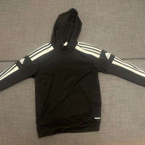 Svart Adidas hoodie med vita ränder - Svart hoodie från Adidas med klassiska vita ränder längs ärmarna och Adidas-logga på båda armarna. Tröjan har huva med snörning och en stor magficka framtill. Tillverkad i mjuk polyester, perfekt för chill eller sport. Snygg och sportig stil!