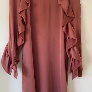 Rosa klänning med volangärmar - Supersöt rosa klänning med långa volangärmar och rak passform. Klänningen har en mjuk, flowy känsla och är perfekt för dig som gillar en feminin och trendig look. Ärmarna har flera lager volanger som ger extra rörelse och stil. Använd ett par gånger.