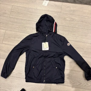 Navy vindjacka från Moncler - Snygg mörkblå vindjacka från Moncler med huva och dragkedja. Jackan har Moncler-logga på ärmen och rödvit rand på huvan. Två fickor framtill med tryckknappar och justerbar nederkant. Perfekt för dig som vill ha en stilren och sportig look. ALDRIG ANVÄND!!!