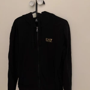 Svart hoodie från EA7 Emporio Armani - Svart hoodie från EA7 Emporio Armani med guldfärgad logga på bröstet. Tröjan har dragkedja framtill, huva med snörning och långa ärmar. Perfekt för en stilren och sportig look.