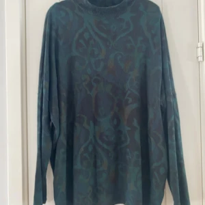 Mörkgrön mönstrad polotröja - Säljer en oversized polotröja med mörkgrönt och blått abstrakt mönster. Tröjan har ribbad hög krage och långa ärmar, perfekt för dig som gillar chill och loose fit. Materialet är stickat och mjukt, passar dig som vill ha något bekvämt och snyggt. Mått Över bröstet: ca 84 cm Längd från axeln och ner: ca 77 cm  74% viskos, 21% polyester och 5% elastan  Kommer från ett djur och rökfritt hem. 