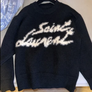 Svart stickad Saint Laurent tröja S - Cool svart stickad tröja från Saint Laurent med vit fuzzy logga framtill. Tröjan har rund hals och långa ärmar, ribbade muddar och en loose passform. Perfekt för dig som gillar statement-plagg och designer vibes. Vid frågor kontakta mig!