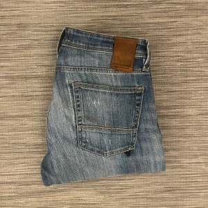 Blå slitna jeans från Jack & Jones - Säljer ett par blå jeans från Jack & Jones med snygga slitningar och hål på knäna. Jeansen har klassisk femficksdesign, bruna detaljer vid linningen och en straight passform. Materialet är robust denim med ljusa tvättade partier och slitna effekter. Finns missfärgningar och slitningar, alltså inte i bästa skick. Storlek: W31/L34