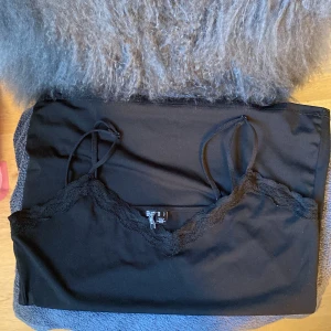 Svart linnetopp med spets från Pieces - Svart linnetopp från Pieces med tunna axelband och snygg spetskant längs urringningen. Toppen är enkel men ändå elegant, perfekt att styla med jeans eller kjol. Mjuk och skön känsla tack vare bomullsmaterialet.