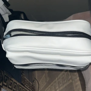 Vit axelväska från Zara - Snygg vit axelväska från Zara med två stora fack och svarta dragkedjor. Väskan har en justerbar axelrem i svart och är tillverkad i slätt syntetmaterial. Perfekt storlek för att få plats med allt du behöver och stilren design som passar till många outfits.