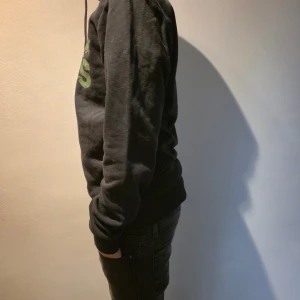 Svart hoodie från Jack & Jones - Svart hoodie från Jack & Jones med grön logga framtill. Klassisk modell med huva och snörning, ribbade muddar vid ärmslut och midja. Perfekt för chill dagar eller när du vill ha en enkel streetstyle-look. Skriv vid funderingar. Jag själv är 165-170 på bilden.