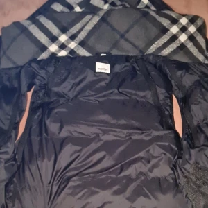 Svart pufferjacka från Burberry - Skaffa dig en svart down jacket från Burberry med klassiskt rutigt foder. Jackan är vadderad och har en modern utseende. Perfekt för kommande kalla dagar när du vill vara både varm och stilren. Pris kan diskuteras vid snabbäffar. Skriv för mer info.