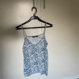 Blåvit vackert och unikt linne - Säljer en snygg linnetopp med tunna axelband och paisley-mönster i blått och vitt. Toppen har en v-ringad front och ärmlös design, perfekt för varma dagar. Materialet är mjukt och lätt, troligen bomull eller polyester. Supersnygg till jeans eller shorts!