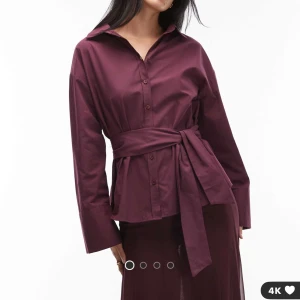 Vinröd skjorta med knytband - Stilren vinröd skjorta med lång ärm, klassisk krage och knappar framtill. Skjortan har ett brett knytband i midjan som ger en snygg siluett. Perfekt för dig som gillar en trendig och elegant look.