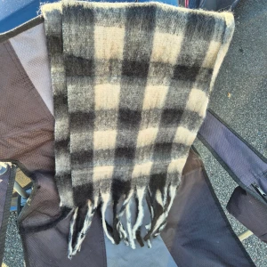Rutig ullhalsduk i svart och beige - Mysig halsduk med rutigt mönster i svart, grått och beige. Den är stickad i ull och har långa fransar i ändarna. Perfekt för att hålla värmen under kyliga dagar och ger en cool, klassisk vibe till din outfit. Helt ny och supermjuk 