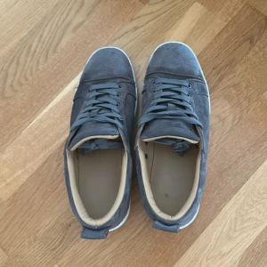 GE PRISFÖRSLAG STORLEK 43 MEN PASSAR 42 - Snygga sneakers från Christian Louboutin i grå mocka med klassisk röd sula och vita kanter. Skorna har rund tå, snörning och beige innersida. Perfekta för dig som vill sticka ut med exklusiva detaljer och streetstyle-vibe.