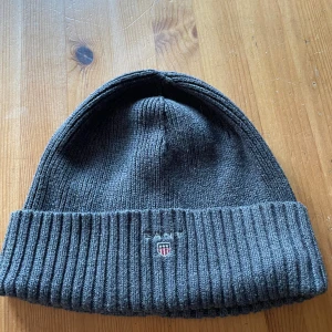 Mörkgrå ribbstickad mössa från GANT - Snygg mörkgrå ribbstickad mössa från GANT med uppvikt kant och diskret broderad logga framtill. Tillverkad i 100% bomull för en mjuk och bekväm känsla. 