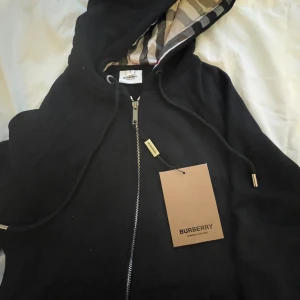 Svart hoodiejacka från Burberry - Svart hoodiejacka från Burberry med dragkedja och klassiskt rutigt foder i huvan. Jackan har snörning i huvan och snygga metalldetaljer på snörena. Perfekt för dig som vill ha en stilren och lyxig look med ikoniska Burberry-detaljer.
