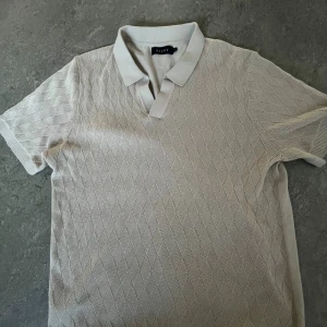 Beige pikétröja från Riley XL - Snygg beige pikétröja från Riley i storlek XL. Tröjan är tillverkad i 100% bomull och har ett diskret diamantmönster framtill, klassisk krage och korta ärmar. Perfekt för en clean och stilren look.Den passar också som L