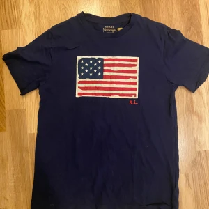 Mörkblå t-shirt🇺🇸 Polo Ralph Lauren - Mörkblå t-shirt från Polo Ralph Lauren med tryck av amerikanska flaggan och RL-broderi på bröstet. Klassisk passform och rund hals. Tillverkad i mjuk bomull, perfekt för en avslappnad stil.