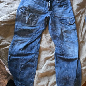 Blå jeans från Gorilla Wear med fickor - Säljer ett par blå jeans från Gorilla Wear med stora broderade loggor och coola detaljer. Byxorna har flera fickor, bland annat en stor ficka med lock på sidan och broderad text. Snygg streetstil och robust känsla, perfekta för dig som gillar unika jeans.