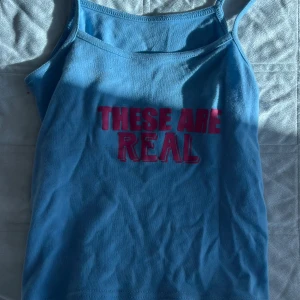 Blå linne med rosa text - Säljer ett blått linne med tunna axelband och trycket 'THESE ARE REAL' i rosa framtill. Perfekt för dig som vill sticka ut med ett statement-plagg. Skön och lätt att bära under varma dagar.