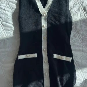 Svart stickad väst/ klänning med pärlknappar - Snygg svart stickad väst/ klänning med vita detaljer och pärlknappar längs hela framsidan. Västen har två dekorativa fickor med vita kanter och pärlknapp. V-ringad och ribbad kant framtill ger en klassisk och chic look.