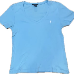 Ljusblå t-shirt från Ralph Lauren - Ljusblå t-shirt från Ralph Lauren med klassisk v-ringning och korta ärmar. Liten vit broderad logga på bröstet. Skön och mjuk bomull som passar perfekt till jeans eller shorts.