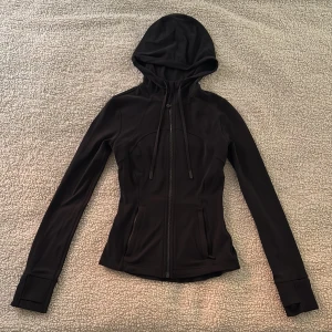 Svart Define jacket från Lululemon - Svart, figursydd hoodiejacka från Lululemon med dragkedja framtill, huva med snörning och långa ärmar med tumhål. Tillverkad i ett stretchigt material (nulu) och med två fickor framtill. Använd endast ett fåtal gånger så i mycket bra skick.