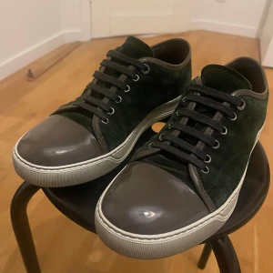 Lanvin sneakers i mörkgrön mocka - Lanvin sneakers med mörkgrön mocka och grå lackad tå. Snygga svarta snören och detaljer i mörkbrunt skinn runt kanten och bak. Platt vit sula med Lanvin-logga undertill. Perfekt för dig som gillar stilrena och lyxiga sneakers.