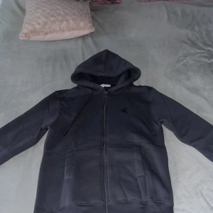 Grå hoodie från Calvin Klein - Grå hoodie från Calvin Klein med dragkedja framtill och broderad CK-logga på bröstet. Tröjan har huva med snörning och stora fickor framtill. Tillverkad i mjuk bomullsmix för extra komfort. Perfekt för en chill och stilren look.St M men passar också lite till S